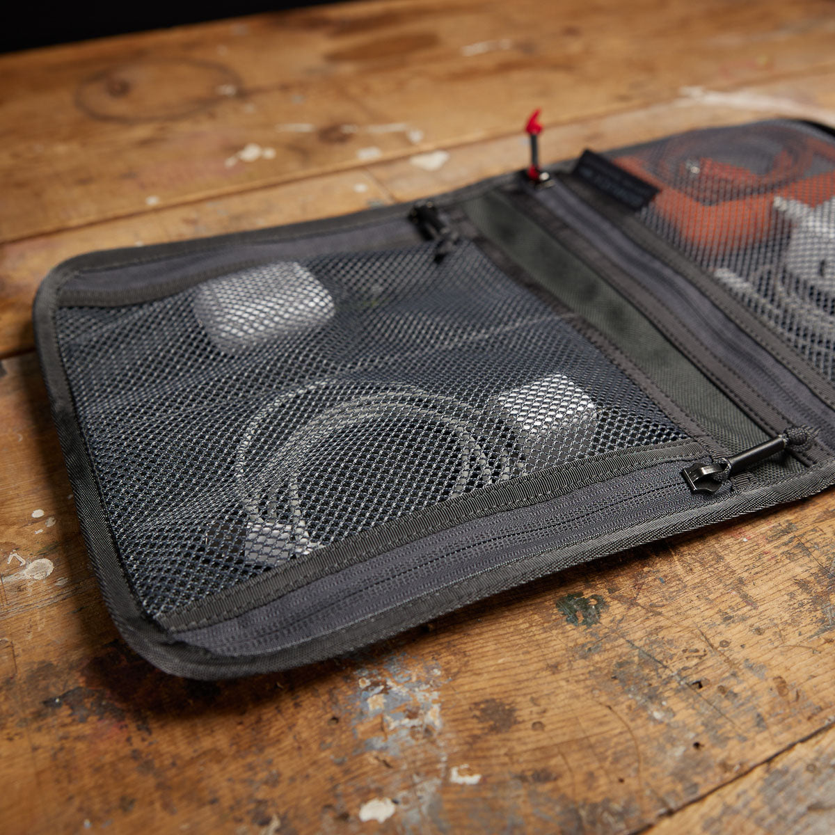 The GORUCK Wire Dopp mesh zippered pouch keeps chargers and cables organized on a worn wooden surface—ideal for managing your travel essentials.