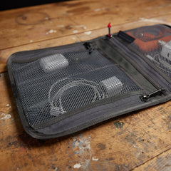 The GORUCK Wire Dopp mesh zippered pouch keeps chargers and cables organized on a worn wooden surface—ideal for managing your travel essentials.