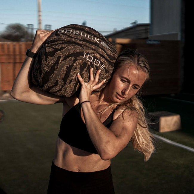 A woman lifts a 100 lb sandbag onto her shoulder outdoors, wearing a black sports bra.