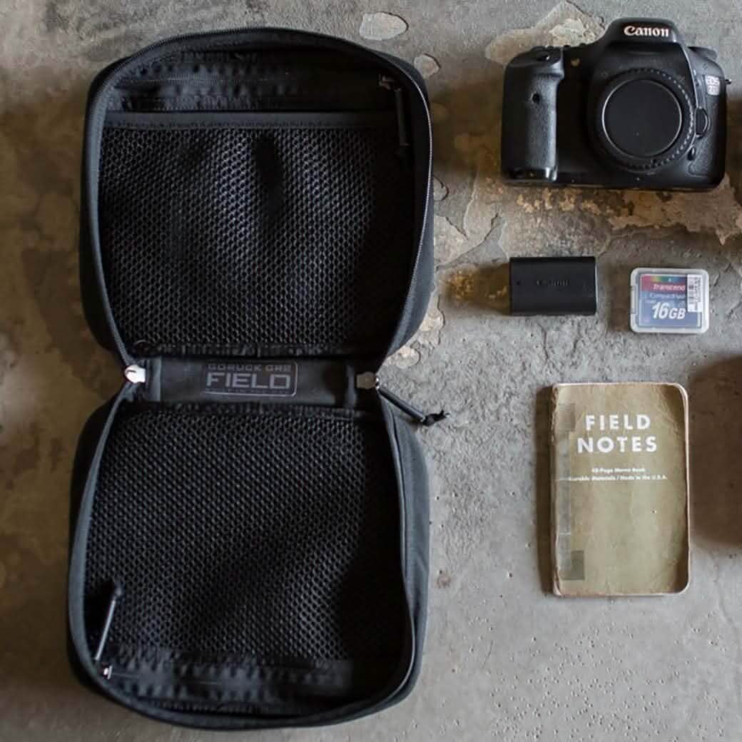 Flat lay of the GORUCK GR2 Field Pocket, a camera, zip pouch, notebook, SD card, battery, and electronics organizer displayed on a textured surface.