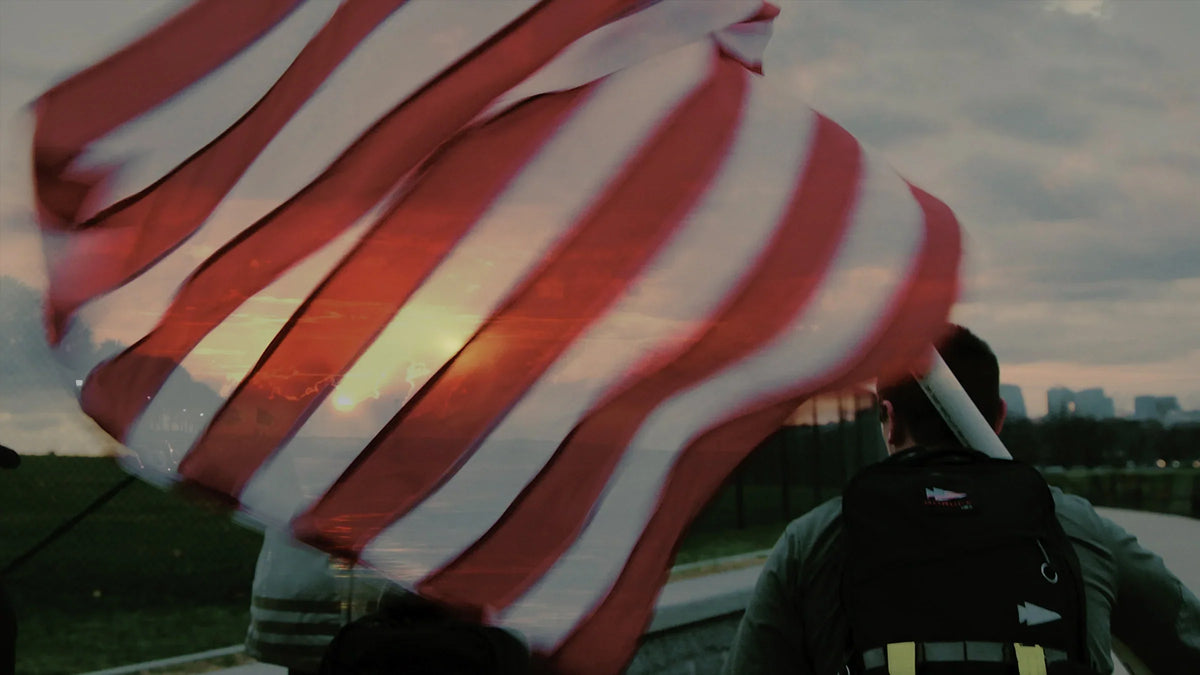 A person with a backpack stands by a large American flag waving at sunset.