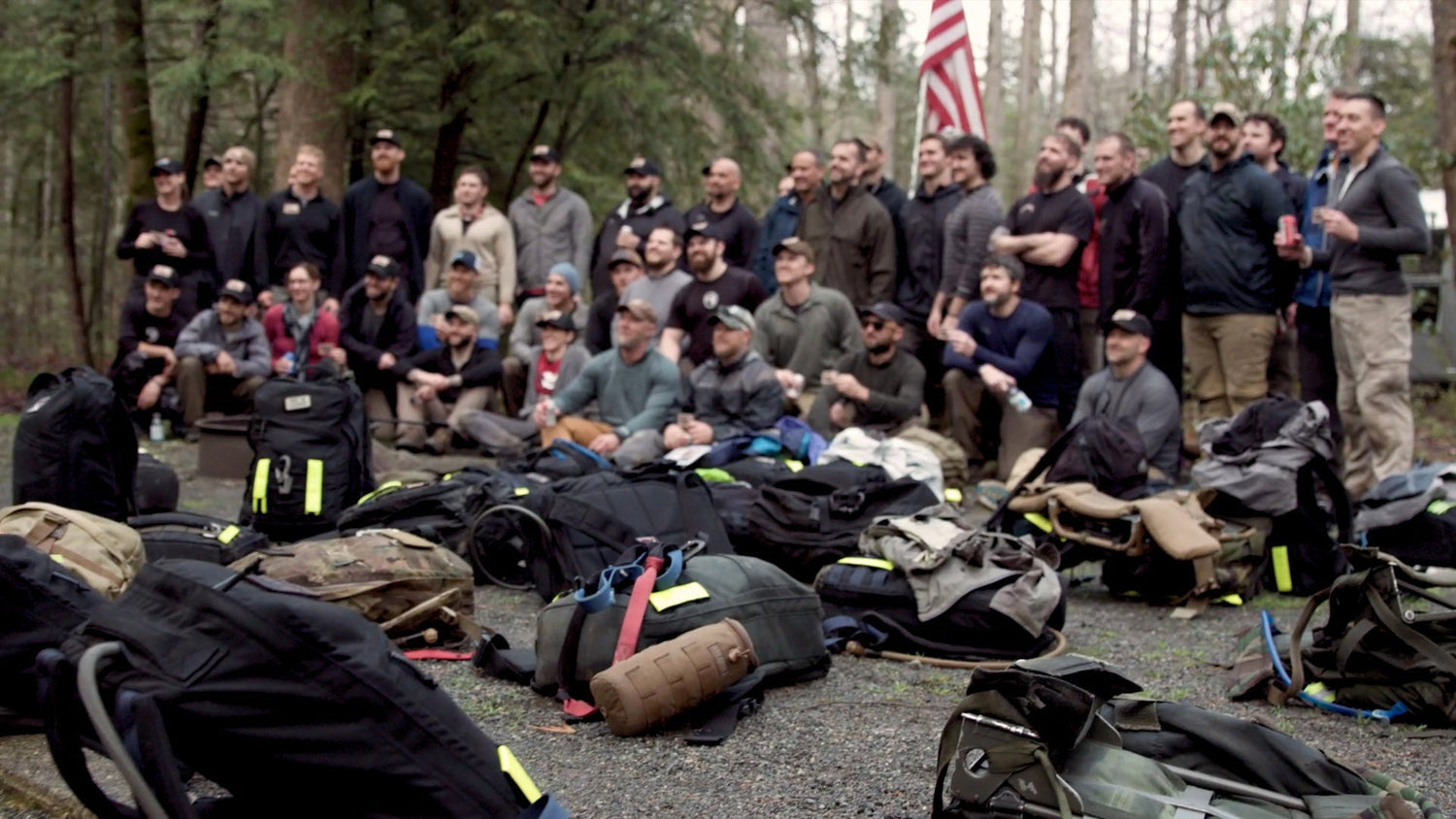 A group of people pose outdoors near trees, with backpacks and gear spread out in the foreground.