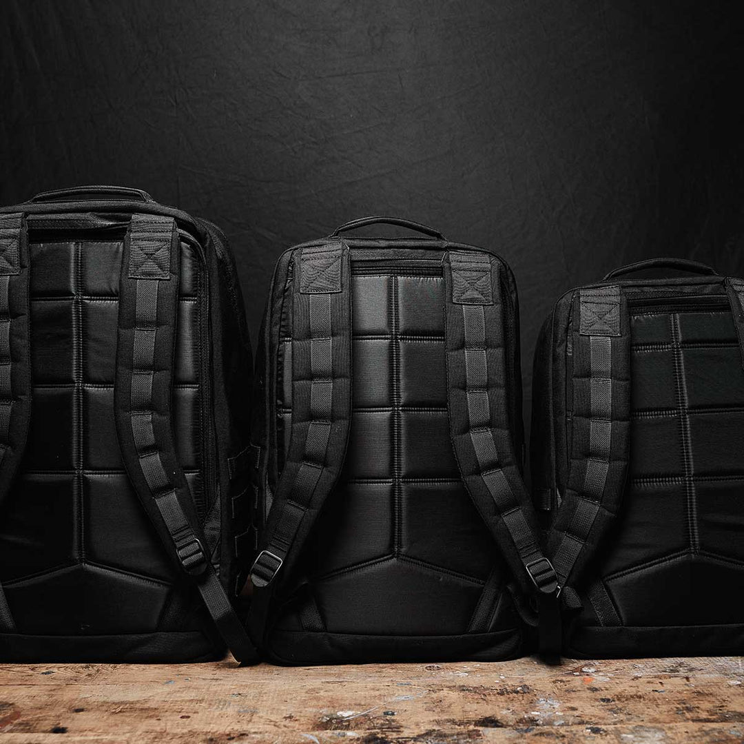 Three black GR2 - USA backpacks by GORUCK, featuring padded straps and crafted from durable 210D CORDURA®, are positioned on a wooden surface against a dark background. Each backpack is carry-on compliant, ensuring a perfect blend of style and convenience for your travel needs.