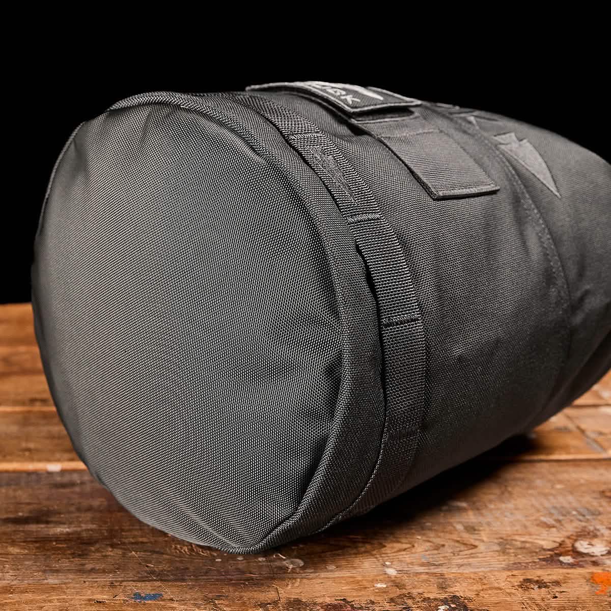 The GORUCK Brick Bag (5-50 LBS), a black cylindrical duffel perfect as a versatile stuff sack for ruck training or use with GORUCK gear, sits on a wooden surface against a black background.