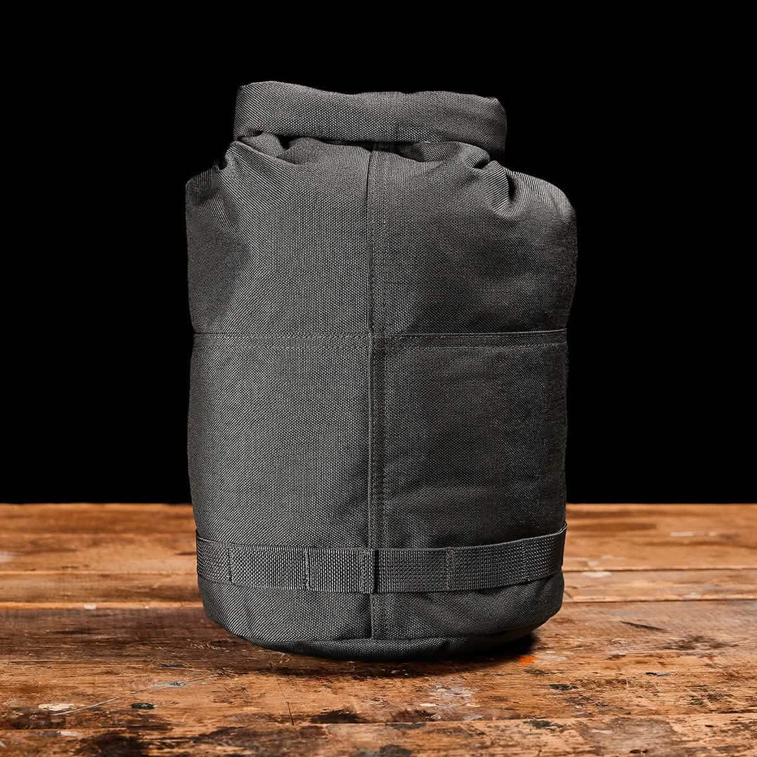 A gray, cylindrical Brick Bag (5-50 LBS) by GORUCK with a rolled top rests on a rustic wooden surface against a black background—a versatile stuff sack ideal for ruck training or pairing with your favorite GORUCK gear.