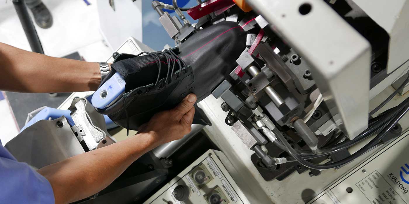 A person operates a machine while assembling a black shoe in a factory setting.