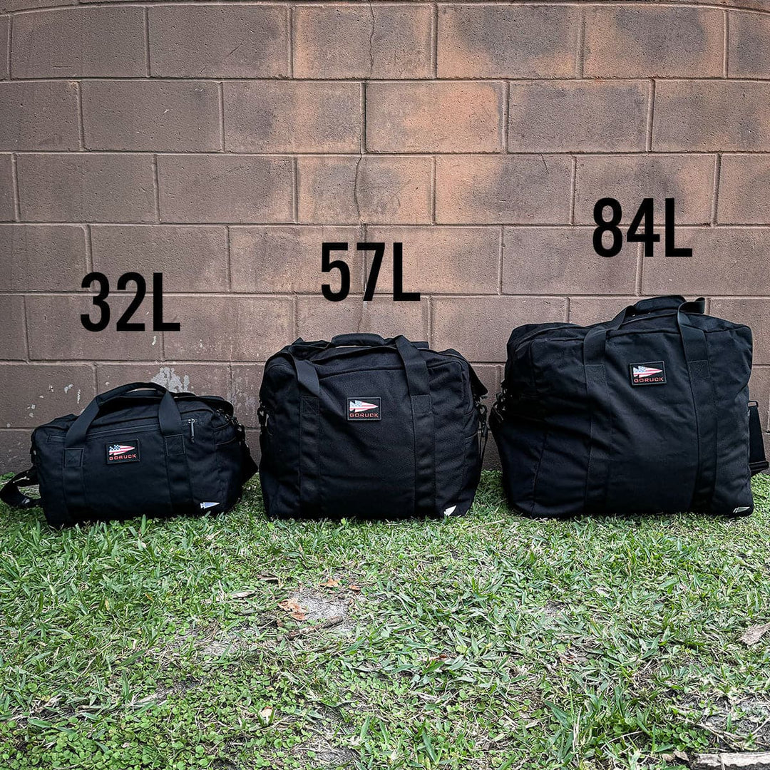 Against a brick wall, three sleek black duffel bags stand in a row, reminiscent of GORUCK's classic Aviator’s Kit Bag. From left to right, they are labeled 32L, 57L, and the impressive Kit Bag - 84L (Includes Shoulder Strap), embodying rugged elegance and adventure readiness.