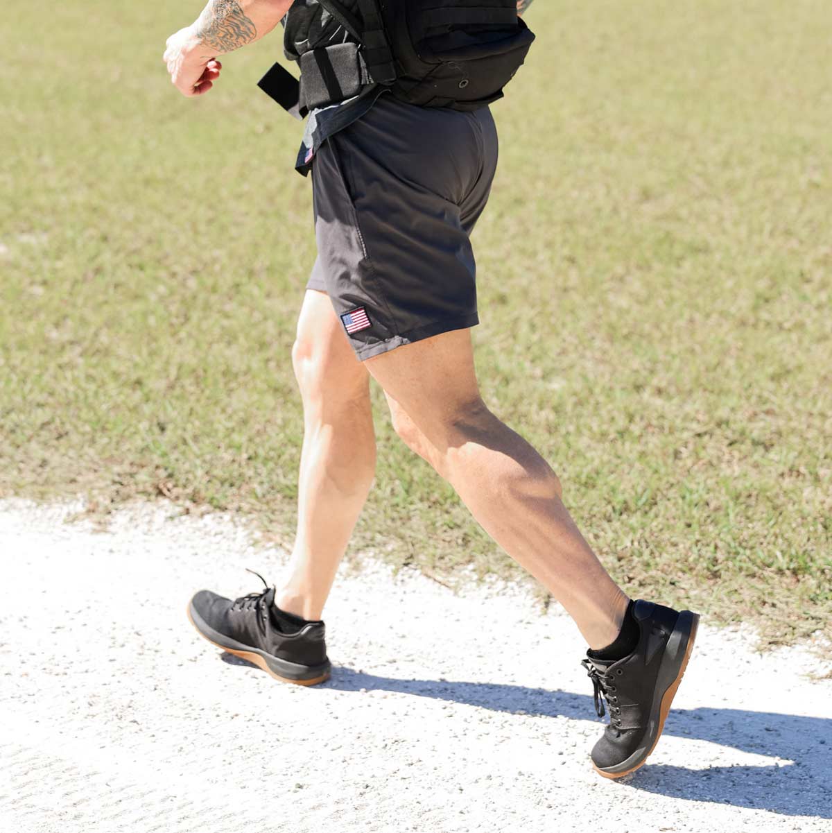 Person walking outdoors on a sunny day, wearing black sneakers, gray shorts, and carrying a backpack.