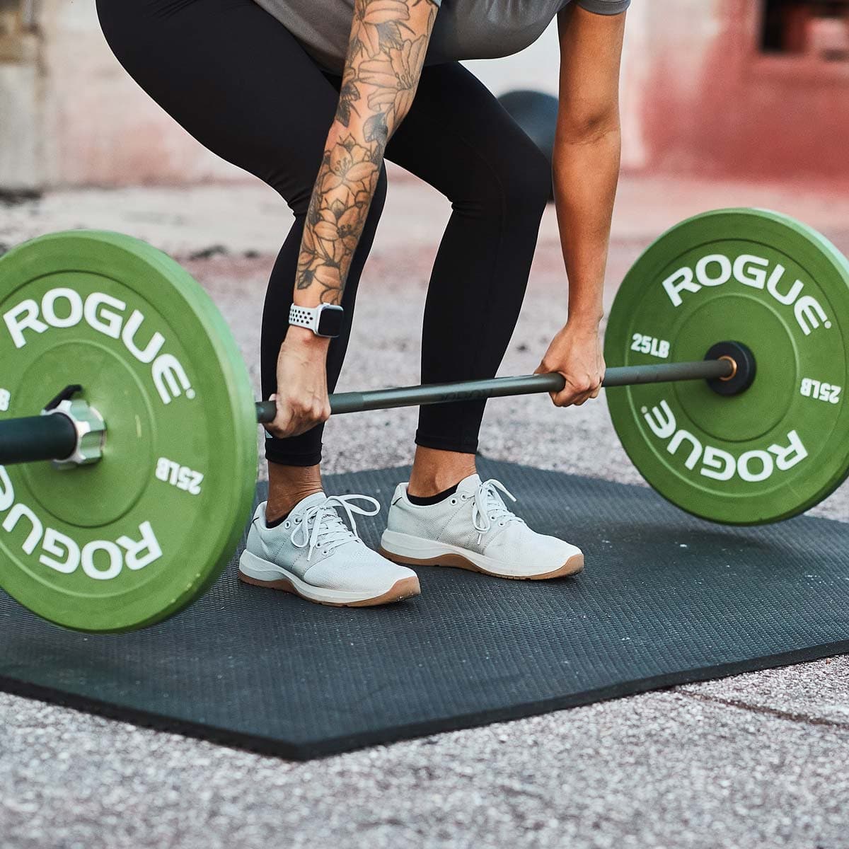 Person with a tattooed arm prepares to lift a barbell with green Rogue weight plates outdoors.