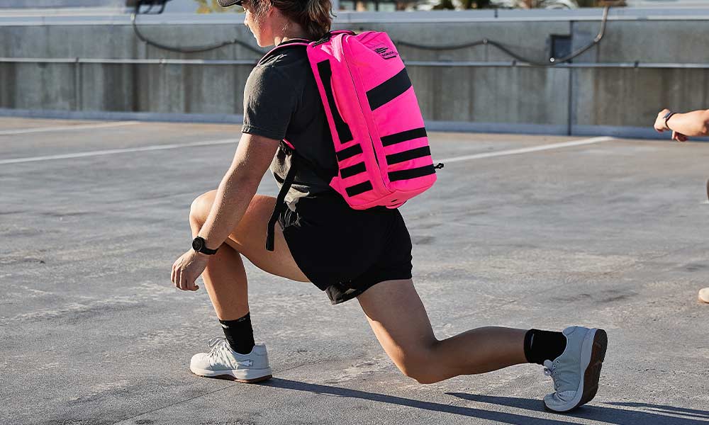 Person wearing a bright pink backpack lunging outdoors on a concrete surface, dressed in athletic wear.