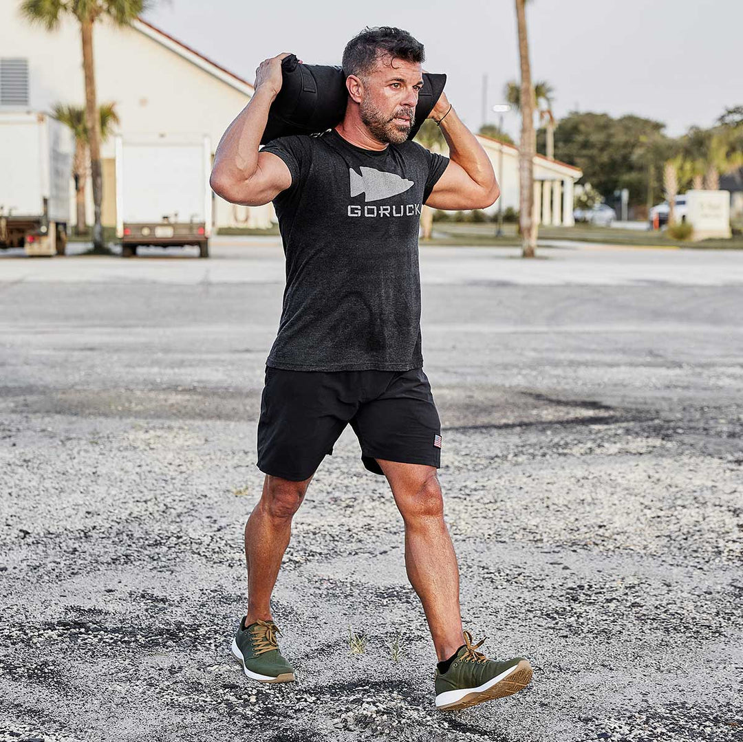 A man walks outdoors in athletic clothing and GORUCK Ballistic Trainers - Green + White + Gum, functional fitness footwear designed for 3X Stability.