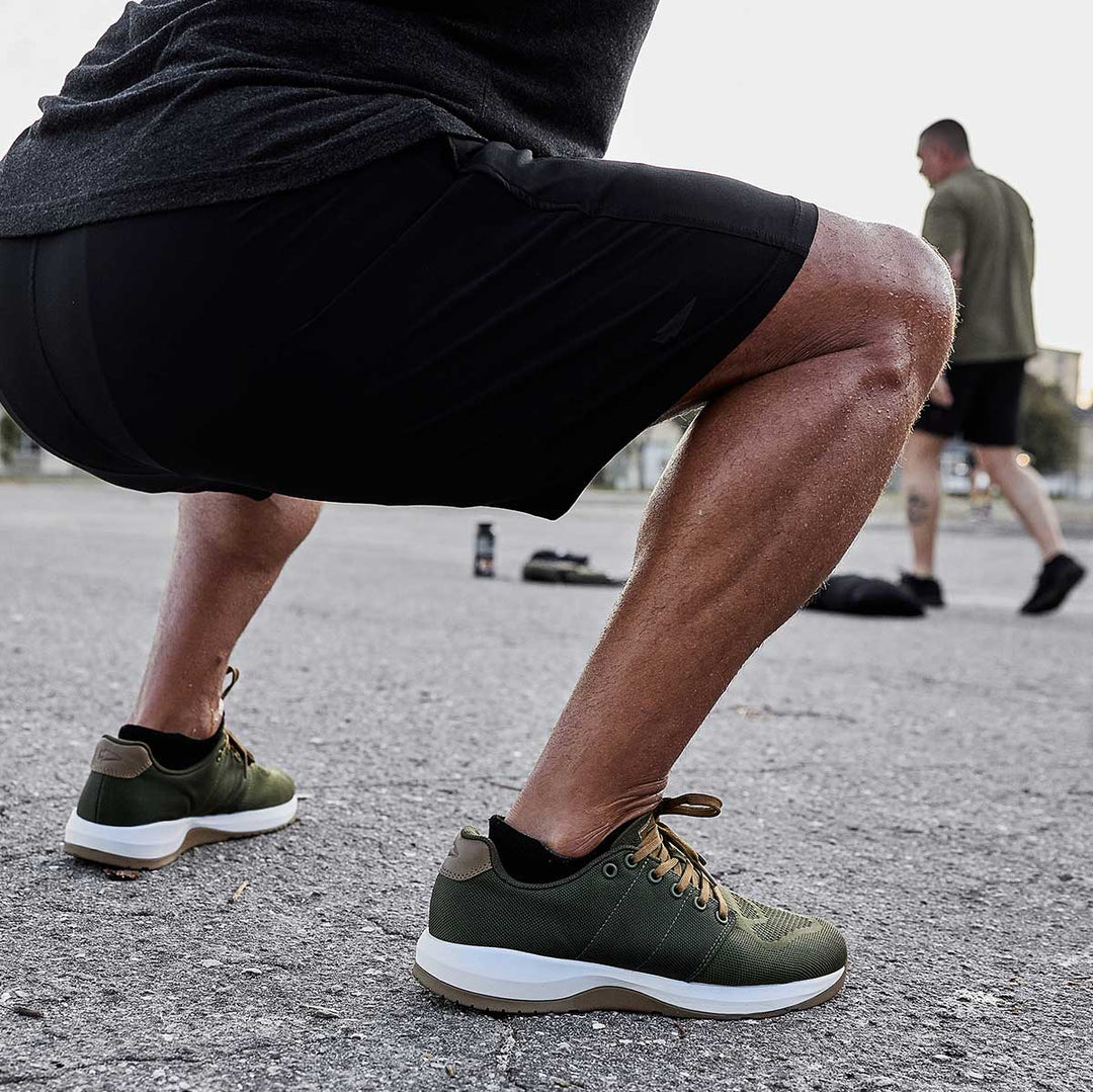 Close-up of a person squatting outdoors in GORUCK Ballistic Trainers - Green + White + Gum athletic shoes, with another individual blurred in the background.