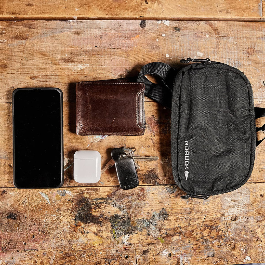 A flatlay on a rustic wooden surface showcases a GORUCK Belt Bag - Ripstop ROBIC® with waterproof zippers, alongside a smartphone, a leather wallet, wireless earbuds in a white case, and a set of keys with a key fob.