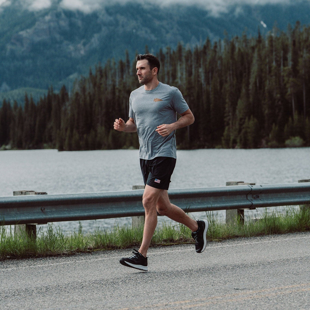 A man, famously called the "Rough Runner," jogs along a road beside a serene lake, surrounded by dense forest and misty mountains. He is outfitted in GORUCK's Men's Rough Runner - Black + White gear, which includes a gray t-shirt, black shorts, and running shoes equipped with a Gradient Density EVA Midsole. The overcast sky adds to the tranquil and natural setting.