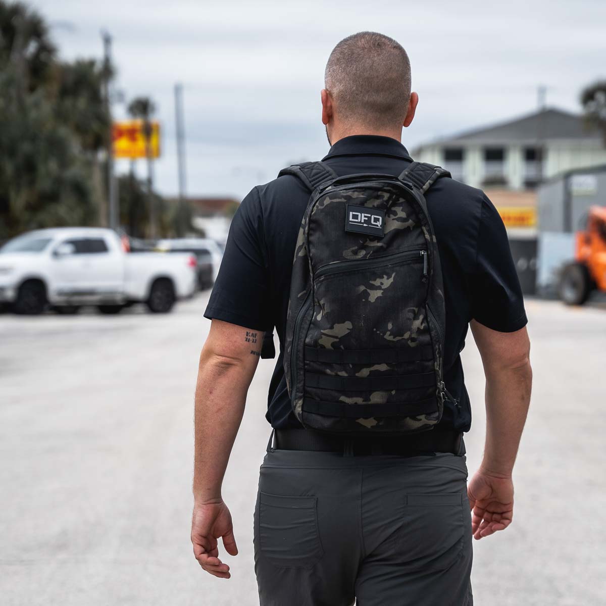 Man with a short haircut wearing a black shirt and camo backpack walks down a street on a cloudy day.