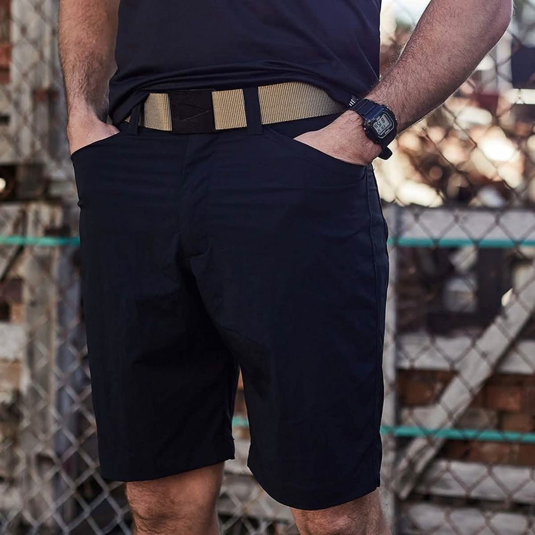 A person stands in front of a chain-link fence, their hands tucked into the pockets of GORUCK's sleek Men’s Simple Shorts made from lightweight ToughDry® fabric, complemented by a beige belt. They sport a black watch while rustic background elements like blurred bricks and wood complete the scene.