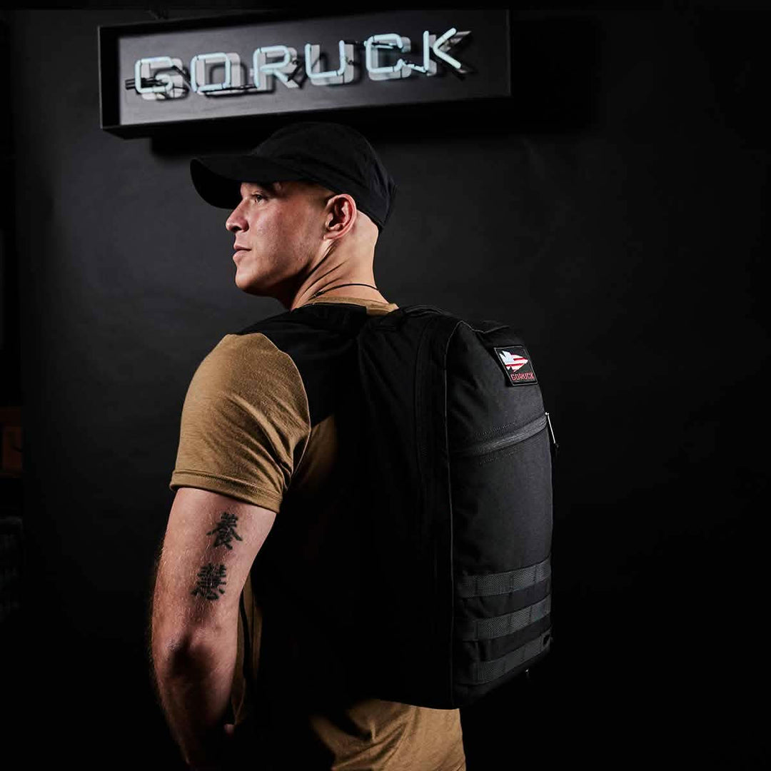 A man in a black cap with a GORUCK Bullet Ruck Laptop - 16L backpack, featuring a laptop compartment, stands in front of a neon "GORUCK" sign.