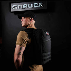 A man in a black cap and tan shirt stands under a lit "GORUCK" sign, wearing the GORUCK Bullet Ruck Laptop - Ballistic Nylon Cordura backpack with a bombproof laptop compartment.