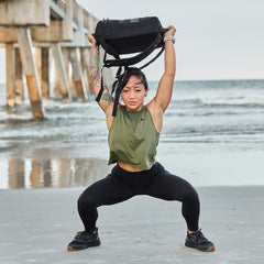 A person is rucking along the shore, lifting a backpack overhead while waves lap at their feet. The reliable Ruck Clubs Starter Kit - Light from GORUCK clings comfortably to their back, beautifully framed by a distant pier and the rolling ocean beyond.