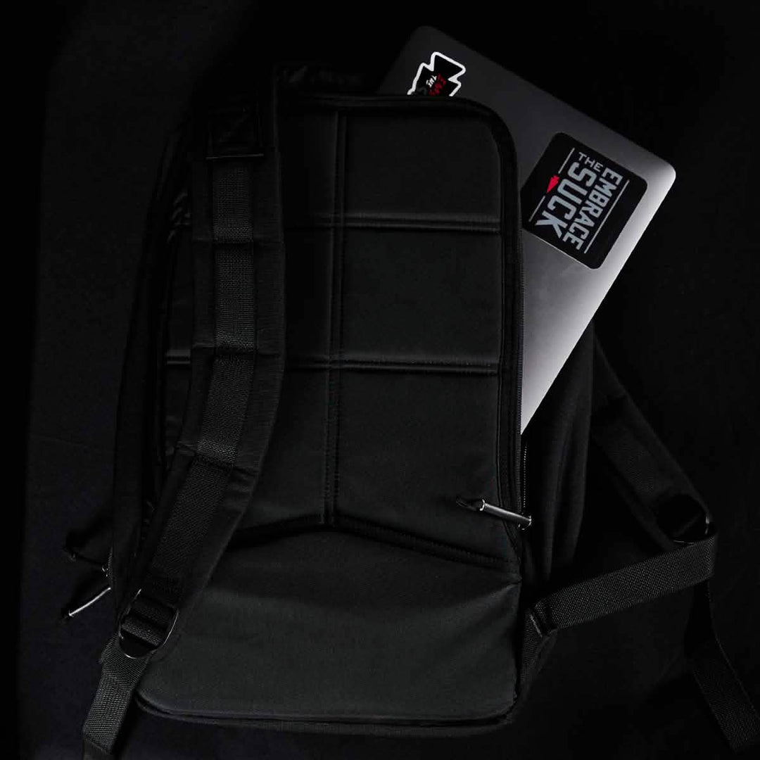 A sticker-covered laptop peeks from the Laptop Compartment of the black GORUCK Bullet Ruck Laptop - 16L backpack, set against a dark background.