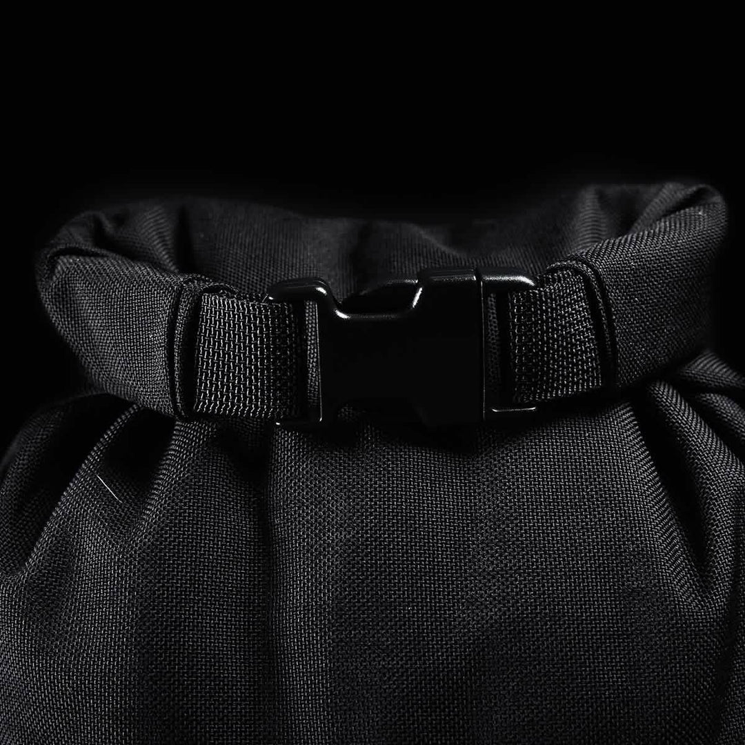 Close-up of a GORUCK Brick Bag (5-50 LBS) in black fabric with a plastic buckle clip, reminiscent of military gear, set against a black background.