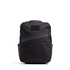 The GORUCK KR1, a sleek black school backpack, features a front diagonal zipper pocket and side pockets while standing upright, offering both style and durability.
