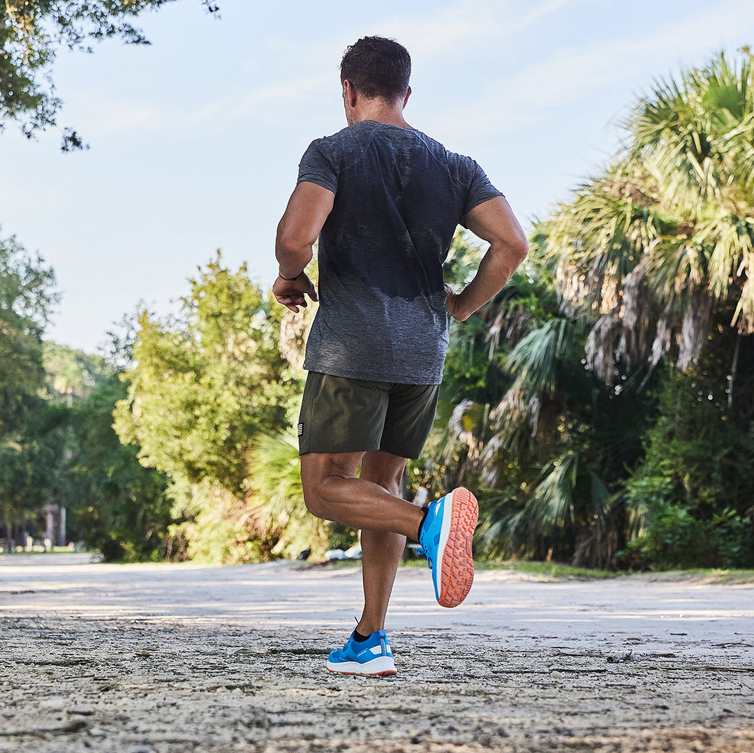 A man jogs on a paved path surrounded by trees and greenery, enjoying the sunny day with ease. His Rough Runner - Electric Blue footwear from GORUCK, equipped with a GRADIENT DENSITY™ EVA MIDSOLE, offers supreme comfort while the versatile road-to-trail outsole adapts seamlessly to any terrain.
