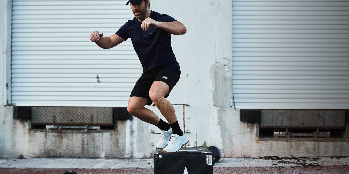 Man in athletic wear jumping onto a black workout box outdoors in front of a white industrial door.