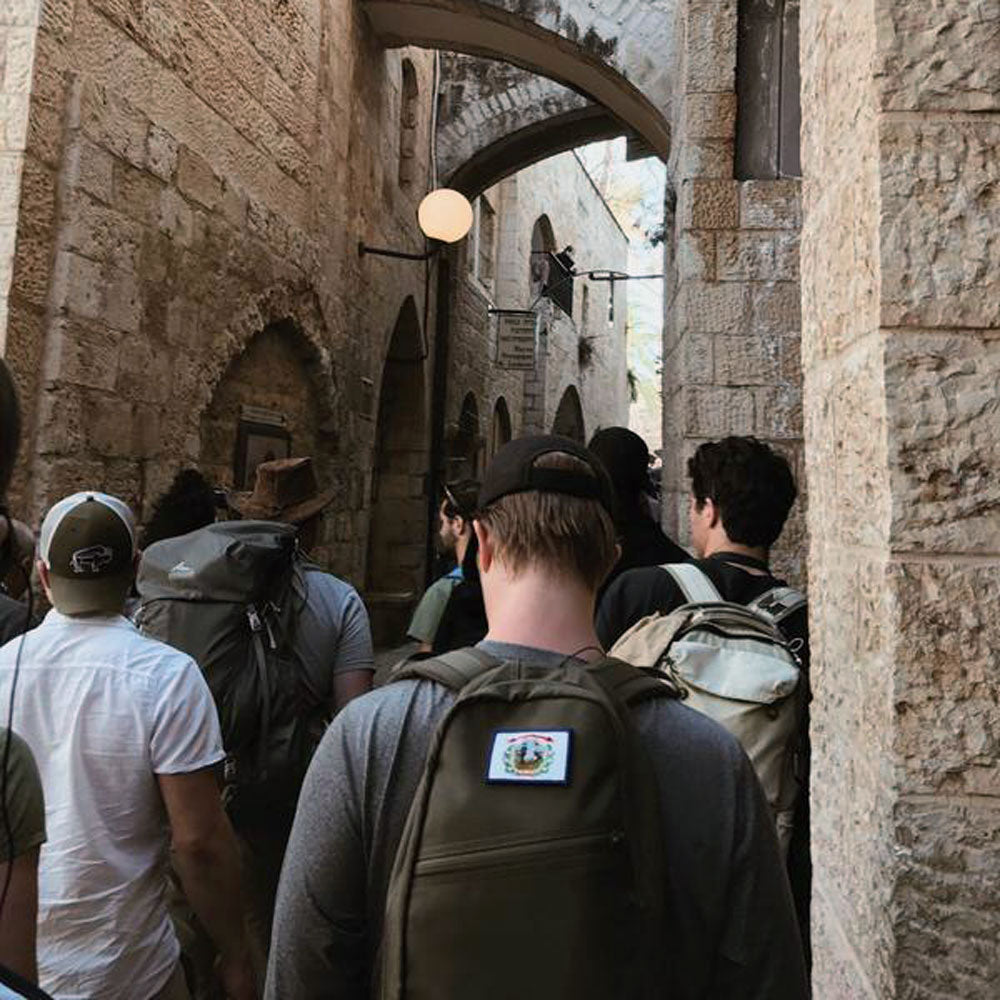 A group of people with backpacks walk through a narrow stone alleyway with arched doorways.