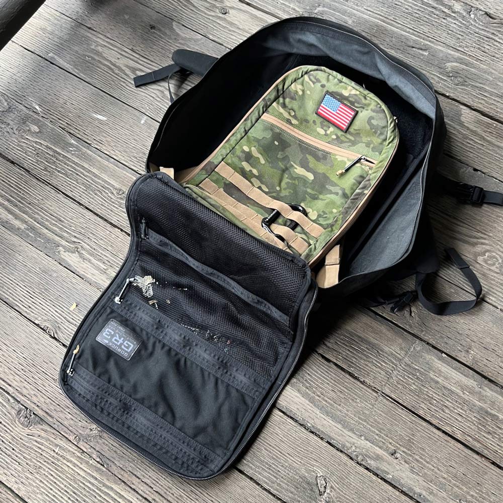 An open black backpack on wood floor, showing a camo insert with a small American flag patch inside.