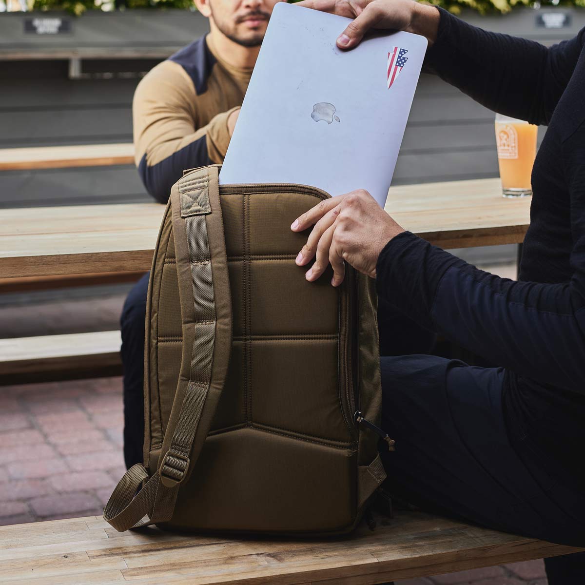 Person placing a silver laptop into a brown backpack while sitting at an outdoor wooden table.