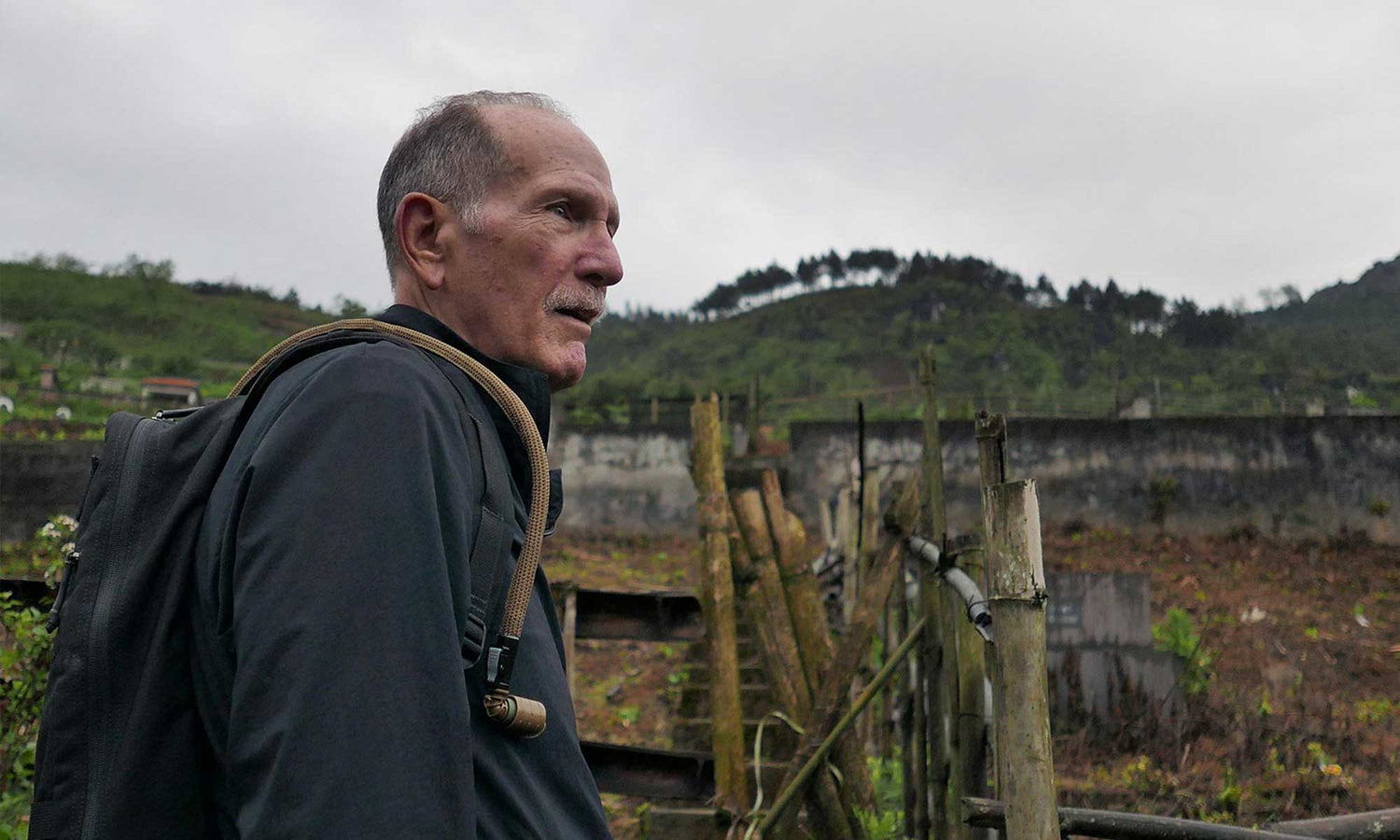 An older man with a backpack stands outdoors near a wooden fence, with green hills in the background.