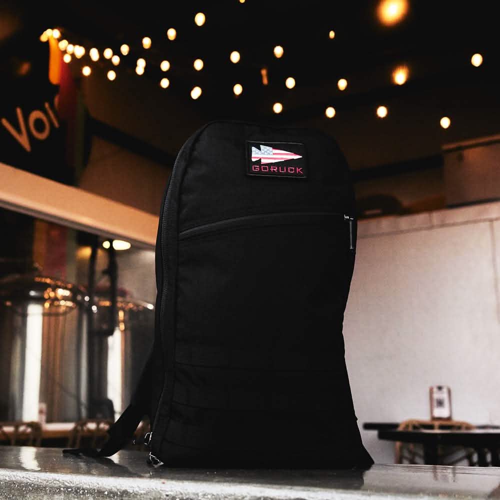 A black GORUCK Bullet Ruck Laptop backpack made from Ballistic Nylon Cordura with a bombproof laptop compartment sits on a counter, string lights glowing in the background.