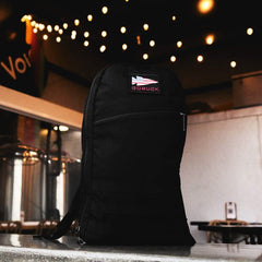 A black GORUCK Bullet Ruck Laptop backpack made from Ballistic Nylon Cordura with a bombproof laptop compartment sits on a counter, string lights glowing in the background.