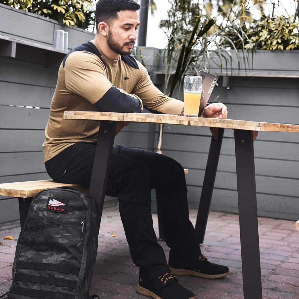 Man sitting at an outdoor table with a drink, looking at his phone, backpack on the ground nearby.
