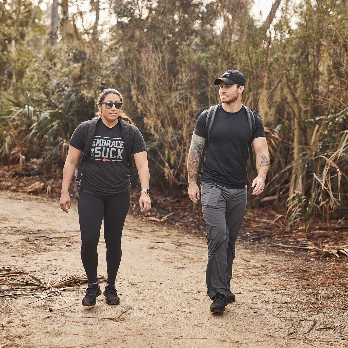 Two people with backpacks walk on a dirt trail through a wooded area, wearing athletic clothing.