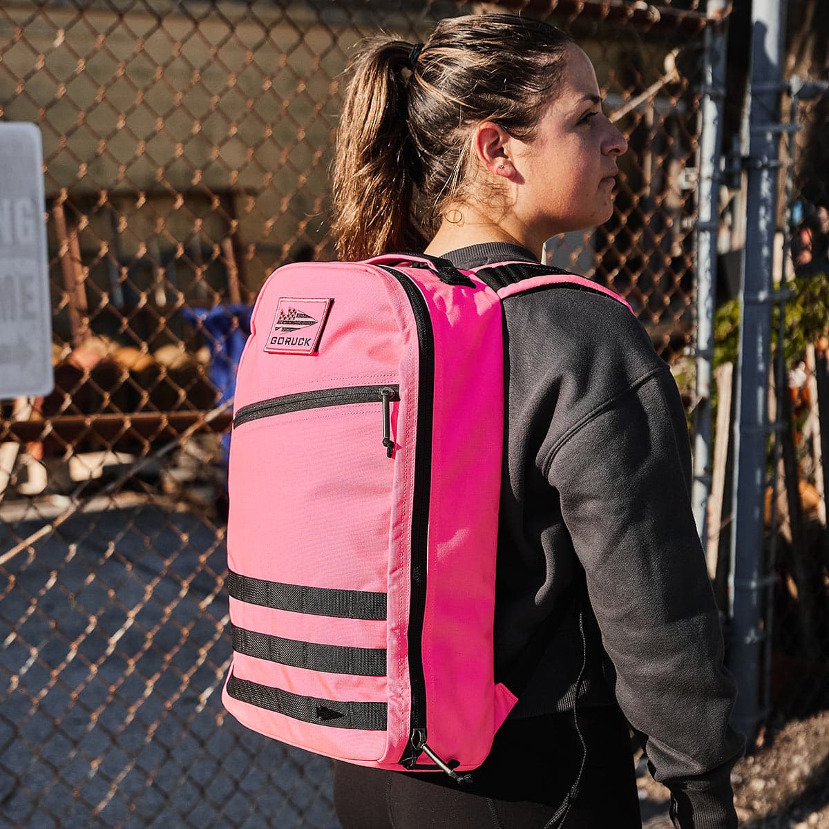 A person outside, sporting a vivid pink 15L Bullet Ruck - Classic by GORUCK, stands near a chain-link fence.