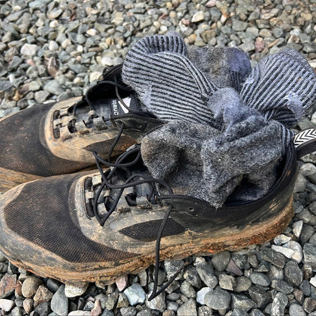 A pair of muddy black sneakers with dirty gray socks stuffed inside, resting on gravel.