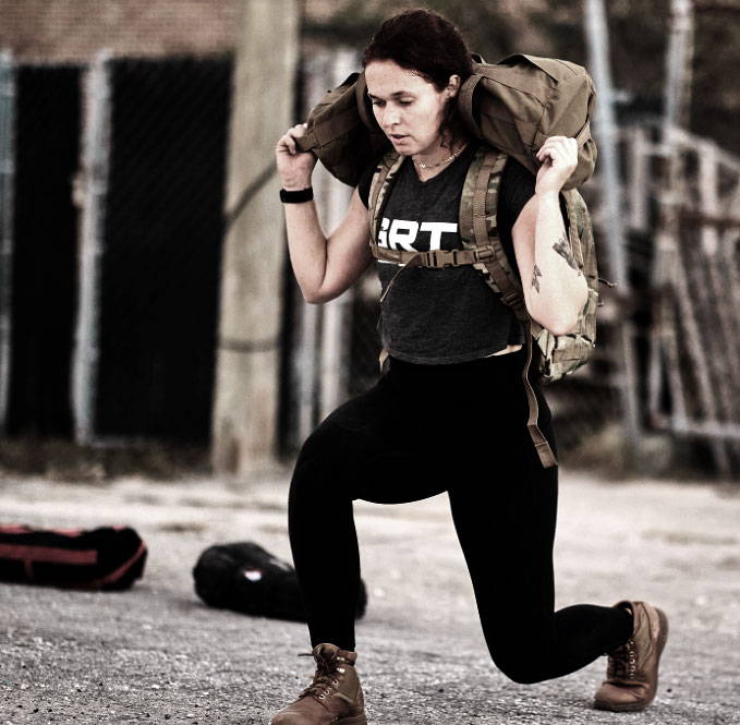 A woman does a lunge outdoors, carrying a heavy sandbag on her shoulders while exercising.
