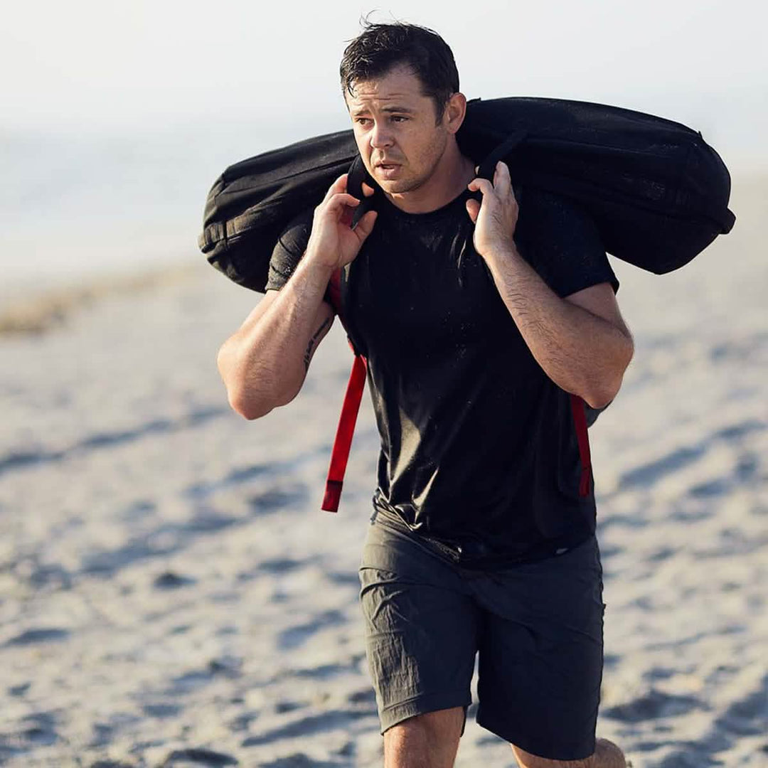 A man, dressed in a black shirt paired with GORUCK's Men’s Simple Shorts made from Lightweight ToughDry® material in gray, carries a large sandbag on his shoulders as he walks along a sandy beach. The background reveals a blurred ocean view beneath a clear sky.