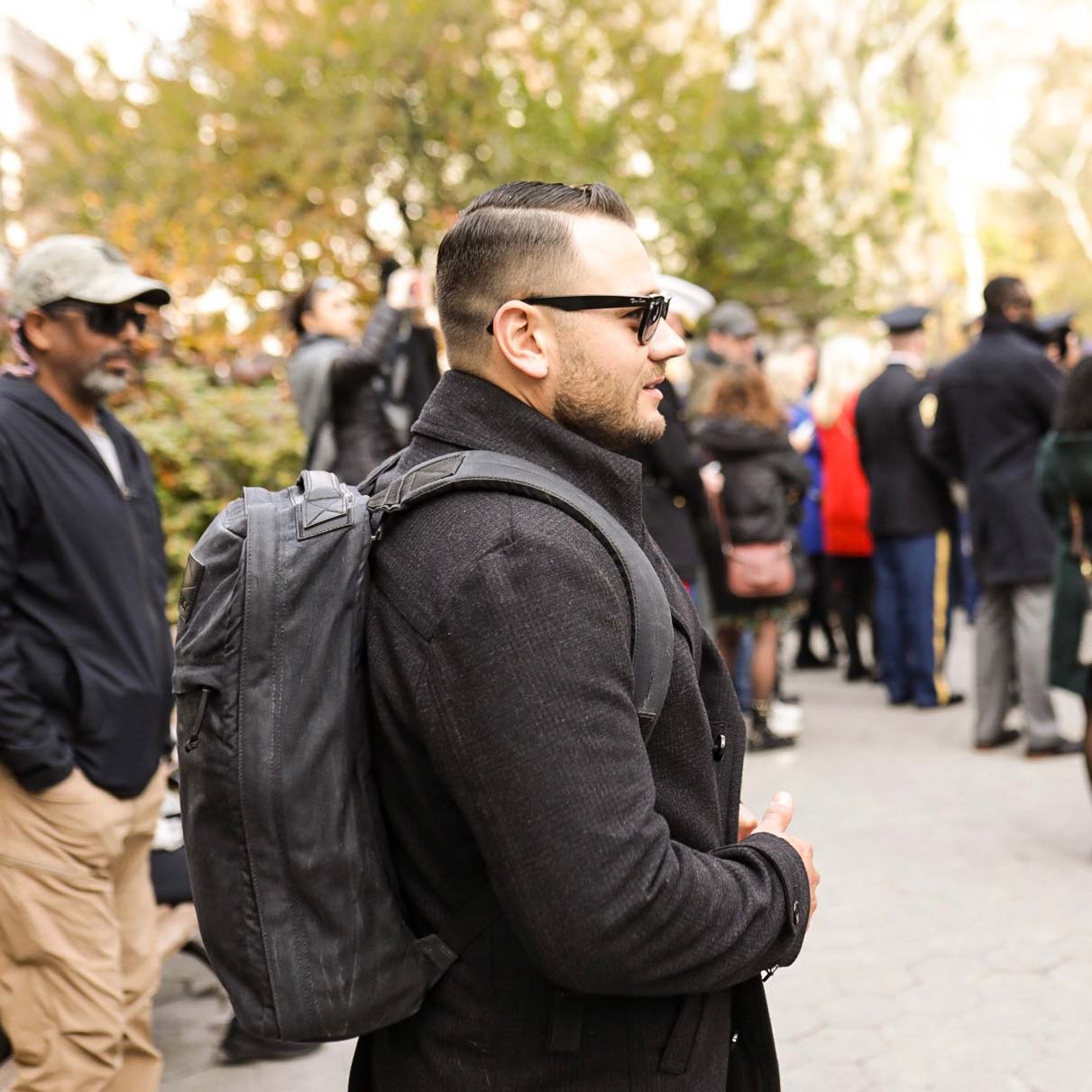 Man wearing sunglasses and a backpack stands outdoors among a crowd of people in a park.