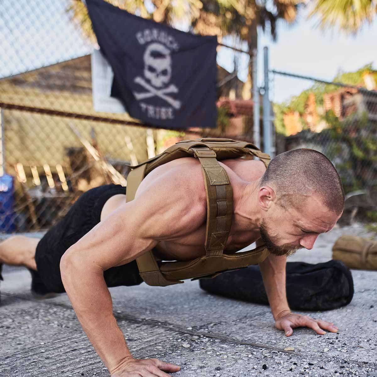 Man wearing a weighted vest does push-ups outdoors near a fence with a skull flag in the background.