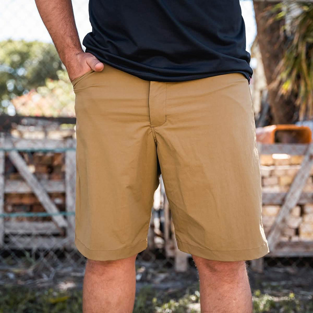 A person is standing outdoors with hands in the pockets of their tan GORUCK Men’s Simple Shorts made from Lightweight ToughDry® material. They are wearing a black t-shirt, while the background shows a wooden fence and trees under a clear sky.