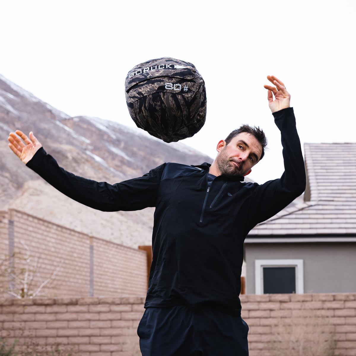 Man tossing an 80 lb GORUCK sandbag outdoors with mountains and a house in the background.