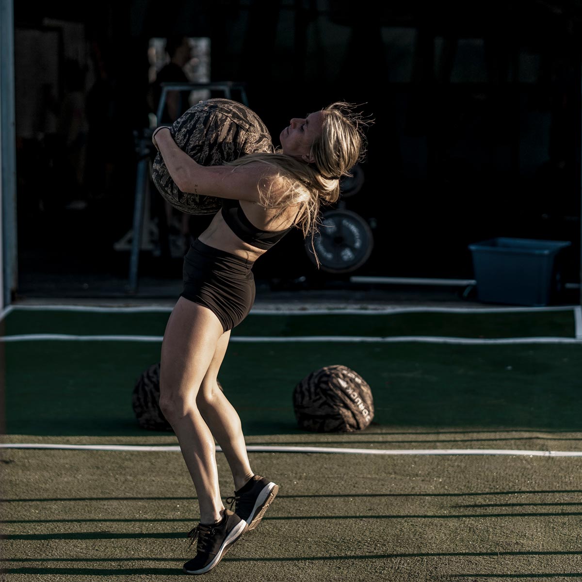 Woman in athletic wear lifts a heavy stone ball outdoors on a green sports court in sunlight.