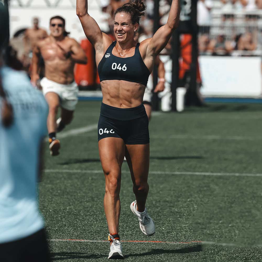 A female athlete, number 046, raises her arms in victory while crossing a finish line on a sports field.
