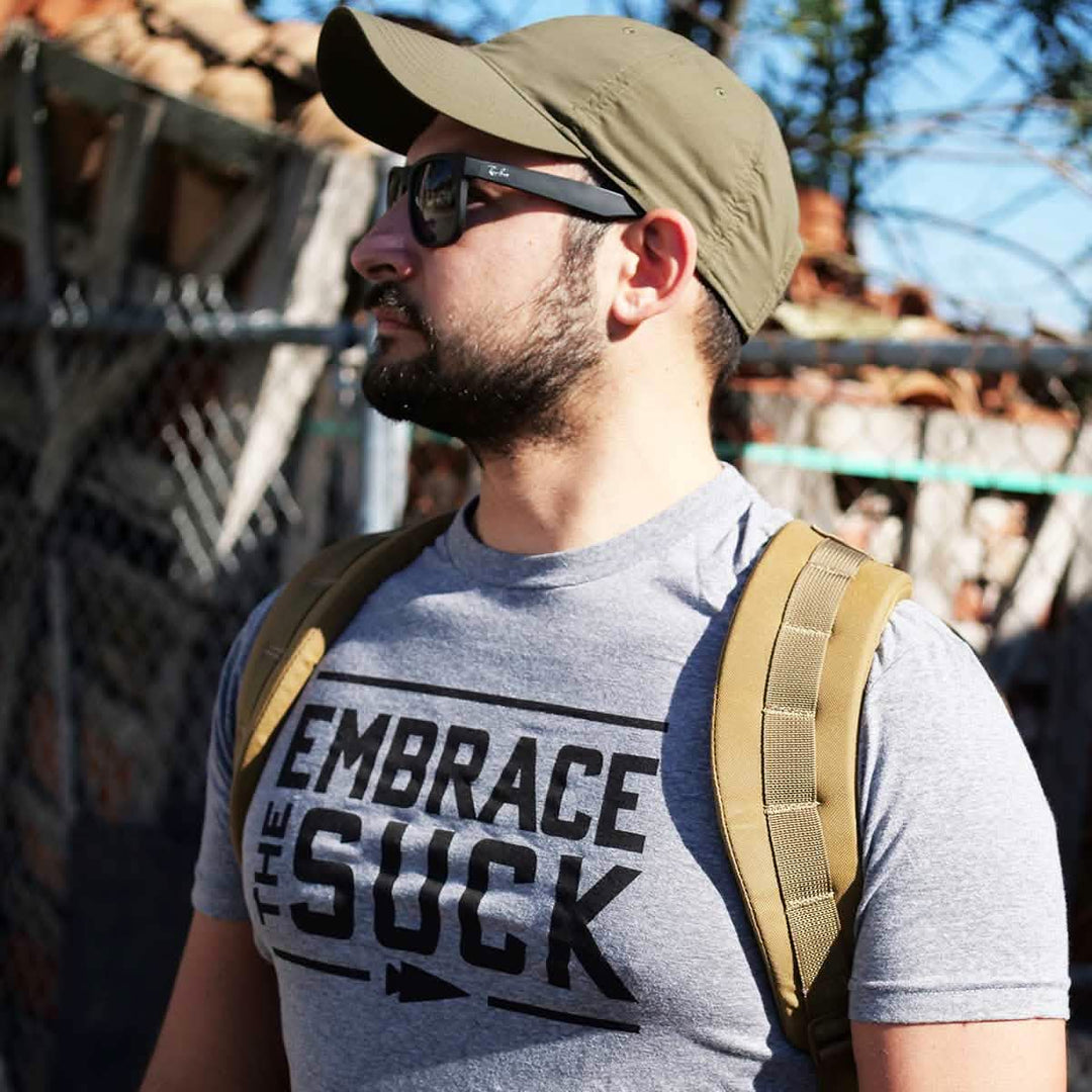 A man in sunglasses and a cap, wearing a backpack and the vendor-unknown Embrace the Suck Tee - Tri-Blend, stands outdoors.
