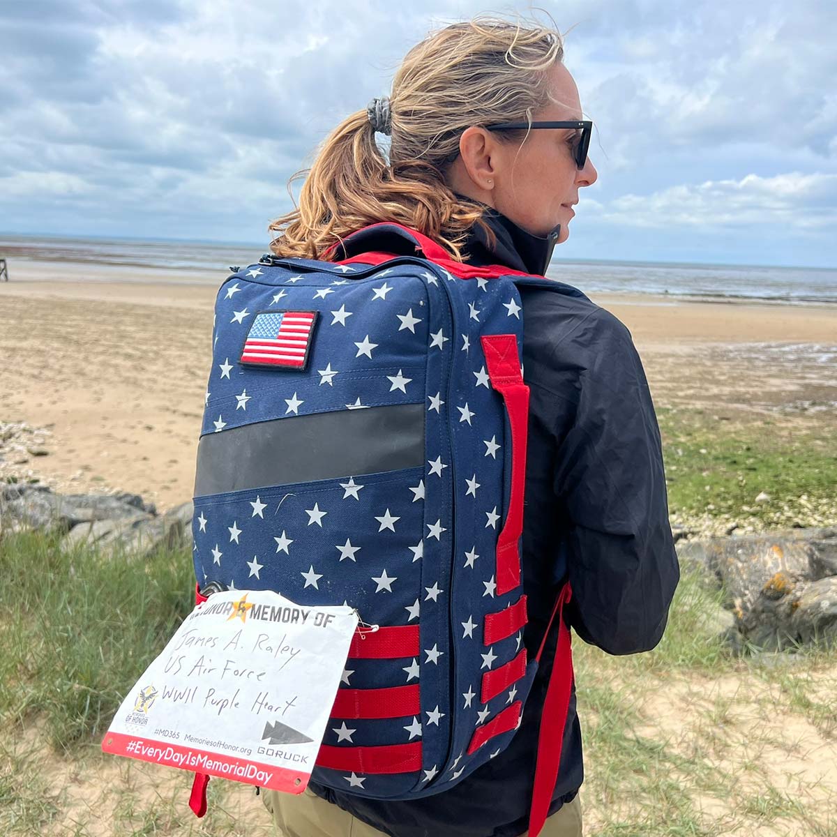 A woman with a star-patterned backpack stands on a beach, facing the ocean.