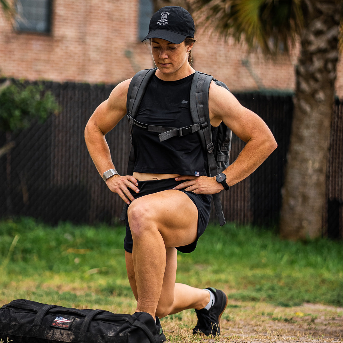 A person in athletic wear and a backpack does a lunge exercise outdoors on grass, next to a duffel bag.