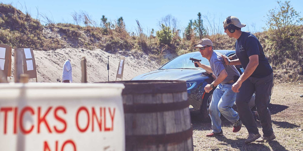 Two men with handguns practice shooting outdoors near a car and barrels, wearing hats and ear protection.